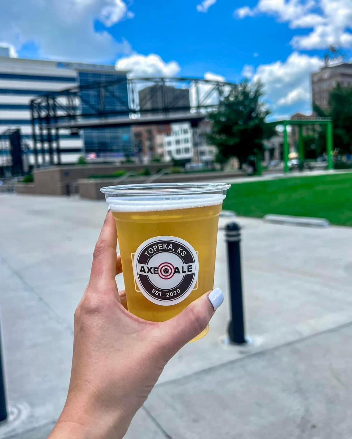 A hand holding a plastic cup of beer with a label from Axe & Ale, set in an outdoor downtown environment with a blue sky and clouds in the background.